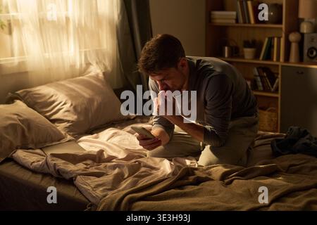 Junger Mann, der nachts mit dem Smartphone auf dem Bett sitzt Stockfoto