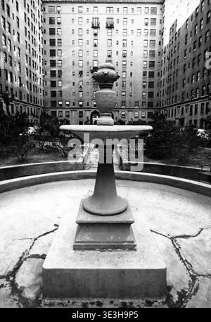 Ein detaillierter Blick aus den 1950er Jahren zeigt einen kunstvollen Brunnen im Innenhof eines Wohnhauses, das sich wahrscheinlich an der Park Avenue und 48th Street in New York City befindet. Die umliegenden mehrstöckigen Backsteingebäude mit zahlreichen Fenstern bilden eine dramatische Kulisse, die an eine urbane Oase erinnert. Dieses Schwarzweiß-Foto von Angelo Rizzuto bietet einen Einblick in die alte New Yorker Architektur und das tägliche Stadtleben. Stockfoto