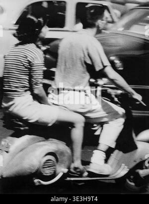 Dieses von Angelo Rizzuto gezeichnete Straßenfoto aus den 1950er Jahren zeigt einen Mann und eine Frau, die einen motorisierten Roller fahren, wahrscheinlich in New York City. Der offene Schuss betont den Alltag und die charakteristische Mode aus der Zeit, mit der Frau in einem gestreiften Oberteil und dem Mann in weißen Shorts. Stockfoto