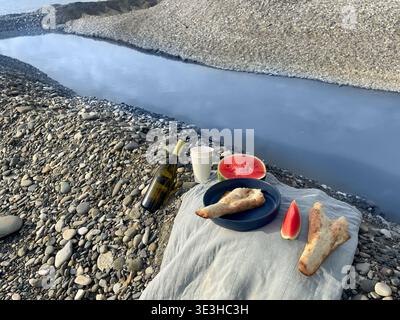Die Menschen versammeln sich am Fluss und teilen sich Essen und Trinken am Kiesstrand. Sie genießen Brot, Wassermelone und Wein, während sie sich unterhalten und entspannen. Stockfoto