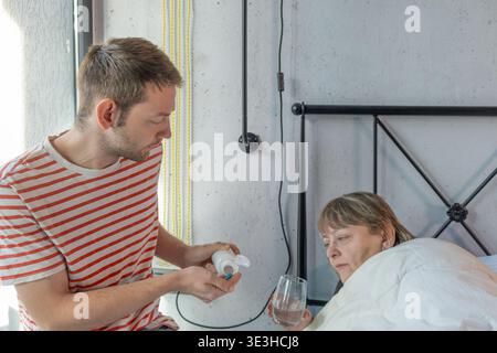 Der Erwachsene Sohn hilft seiner kranken Mutter im Bett, indem er zu Hause Medizin und Wasser gibt. Familiäre Betreuung älterer Eltern, Unterstützung und Verantwortung während der Zeit Stockfoto
