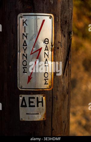 Ein alter Holzmast auf dem Land mit einer Metallplatte, die mit einer griechischen Warnung über Elektrizität befestigt ist, in Korfu Griechenland Stockfoto