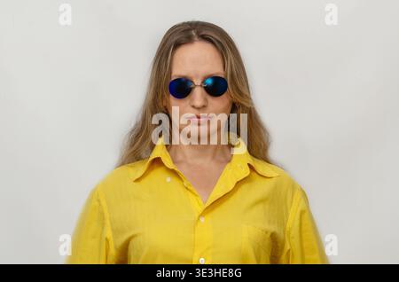 Ernste Frau mit langen Haaren in gelbem Hemd und dunkelblauer Brille, die auf hellem Studiohintergrund posiert. Selbstvertrauen und Individualitätskonzept. Stockfoto