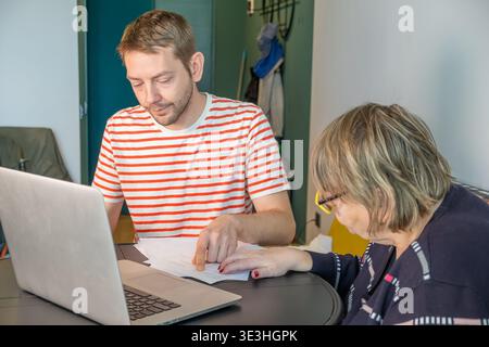 Erwachsener Sohn hilft seiner älteren Mutter, Informationen aus Papierdokumenten in den Laptop einzugeben. Digitale Unterstützung, generationenübergreifendes Lernen und Alltag Stockfoto