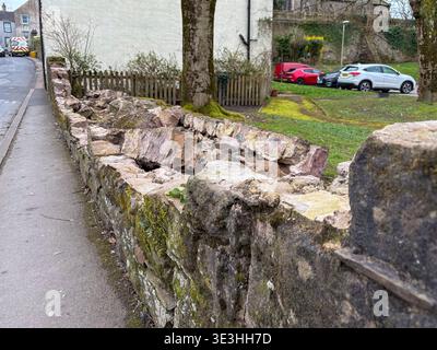 Eine Steinmauer ist auf einer Straße in einem Dorf eingestürzt. Die Wand besteht aus großen, rauen Steinen und ist mit Moos bedeckt. Die Straße ist nass und es gibt s Stockfoto