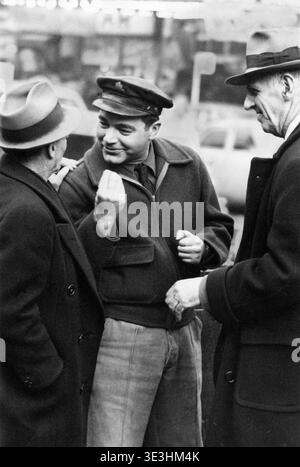 Dieses fesselnde Schwarz-weiß-Straßenfoto aus den 1940er Jahren, aufgenommen von Angelo Rizzuto, zeigt drei Männer, die sich in animierten Gesprächen auf dem Times Square in New York City engagieren. Die zentrale Figur trägt eine unverwechselbare Mütze und Jacke und spricht energisch mit seinen Gefährten, die in Fedoras und Mänteln gekleidet sind und die Alltagsmode der Epoche widerspiegeln. Stockfoto