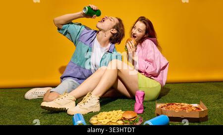 Junger Mann, der aus der Dose trinkt, während die Frau Burger isst und auf grasgelbem Hintergrund sitzt. Stockfoto