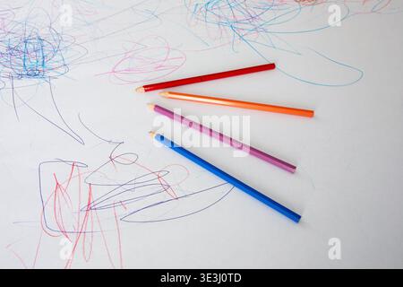 Bunte, ungeschärfte Holzstifte liegen auf einer weißen Papieroberfläche, die mit Kinderkritzeln und Zeichnungen bedeckt ist Stockfoto