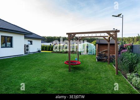 Üppig grüner Hinterhof mit Trampolin-Schaukel, kleinem Gewächshaus, Holzschuppen und blühenden Blumenbeeten Stockfoto