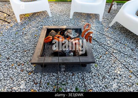 Würstchen, die auf Spießen über einer Metallfeuerkammer mit brennendem Holz und Glut kochen Stockfoto