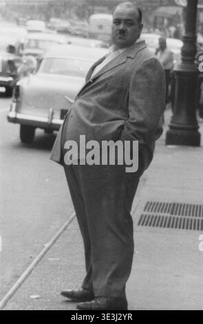 Dieses auffällige Schwarz-weiß-Straßenfoto aus den 1950er Jahren, das wahrscheinlich von Angelo Rizzuto aufgenommen wurde, zeigt einen nicht identifizierten Mann in einem Anzug mit den Händen in den Taschen, der auf einer New Yorker Straße steht. Seine Kleidung und Haltung geben einen Einblick in den Alltag der Mode und des urbanen Lebens dieser Zeit. Stockfoto