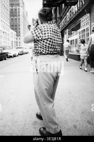 Dieses Straßenfoto, das Mitte der 1950er Jahre vom Fotografen Angelo Rizzuto aufgenommen wurde, zeigt eine Person von hinten, die eine Kamera in der Hand hält und scheinbar ein Bild von einer belebten New Yorker Straßenlandschaft macht. Die Szene umfasst Fußgänger und geparkte Autos mit Blick auf ein Gebäude, das das Schild des Kaufhauses Macy im Hintergrund zeigt. Stockfoto