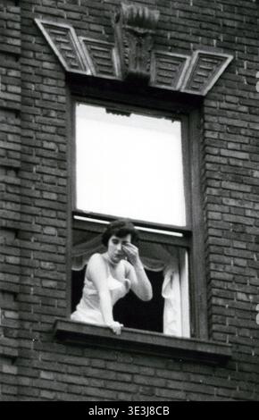 Ein offenes Straßenfoto zeigt eine Frau, die sich wahrscheinlich das Auge abwischt und sich aus einem Fenster in einem alten New Yorker Backsteinhaus lehnt, möglicherweise von Angelo Rizzuto, um die 1950er Jahre Dieses monochrome Bild spiegelt den Alltag und den architektonischen Stil der Epoche der Stadt wider. Stockfoto