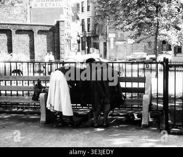 Dieses alte Straßenfoto, das Angelo Rizzuto in den 1950er Jahren in New York City aufgenommen hat, zeigt zwei Frauen, die sich über eine Person lehnen, die auf einer Parkbank liegt. Die Szene fängt einen Moment menschlicher Interaktion und Besorgnis in der urbanen Landschaft ein, charakteristisch für Rizzutos Arbeit, die den Alltag dokumentiert. Stockfoto