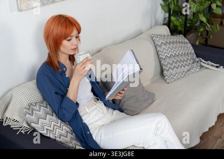 Eine junge Frau in einem blauen Hemd entspannt sich mit einem Notizbuch und trinkt in einem modernen Zuhause. Stockfoto