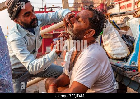 Ein Straßenfriseur rasiert den Bart des Kunden mit einem Rasierer auf den belebten Stufen eines Gheats am Fluss in Varanasi sorgfältig. Varanasi Uttar Stockfoto