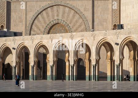 Sonnenbeschienene Fassade der Hassan II Moschee in Casablanca, Marokko, mit sich wiederholenden Steinbögen, komplizierten geometrischen Ornamenten und einigen Besuchern Stockfoto