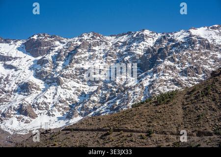 Schneebedeckte Gipfel des Hohen Atlasgebirges in Marokko ragen über felsige Hänge unter einem klaren blauen Himmel auf und bieten eine zerklüftete, malerische Landschaft, die ideal für jeden ist Stockfoto