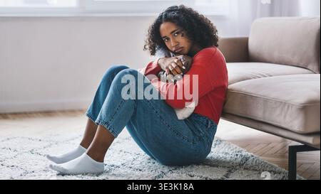 Frau, die auf dem Boden im Wohnzimmer sitzt, mit einem nachdenklichen Ausdruck während der Nachmittagszeit Stockfoto