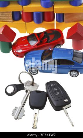 Blick von oben auf Türschlüssel, Fahrzeugschlüssel, zwei Automodelle und ein Spielzeughaus aus Holzblöcken, vor weißem Hintergrund Stockfoto