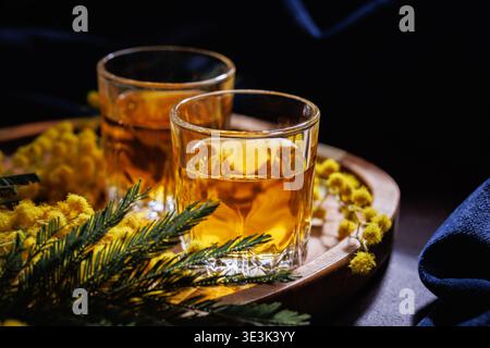 Zwei Gläser Whiskey oder Brandy mit Mimosa-Blumen auf Holztablett elegantes Frühlingsgetränk Stillleben Stockfoto