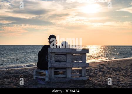 Silhouetten eines Mannes und einer alten Frau sitzen bei Sonnenuntergang auf einer Holzbank am Sandstrand. Familienurlaub, Sonnenuntergang am Strand, Küstenlandschaft. Stockfoto