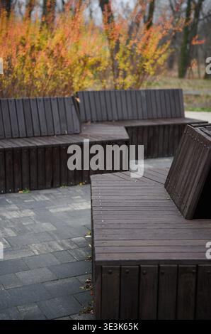 Moderne dunkle Bänke im Herbstpark. Moderner Sitzbereich im Freien mit modularen Holzbänken auf einem Steinpflaster, Herbstlaub hinten. Stockfoto