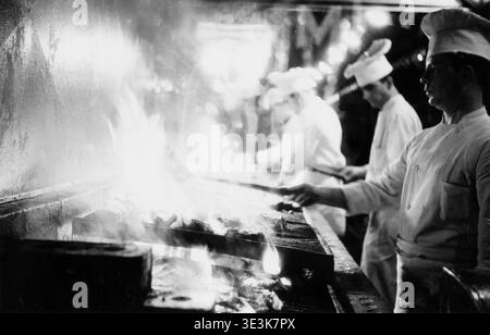 Dieses Vintage-Schwarz-weiß-Foto, wahrscheinlich aus den 1940er-1950er-Jahren von Angelo Rizzuto, zeigt Line-Köche, die in einer belebten Restaurantküche in New York gegrillt haben. Die Szene zeigt die dynamische und intensive Atmosphäre der kulinarischen Arbeit während dieser Zeit und zeigt den Alltag hinter den Kulissen. Stockfoto