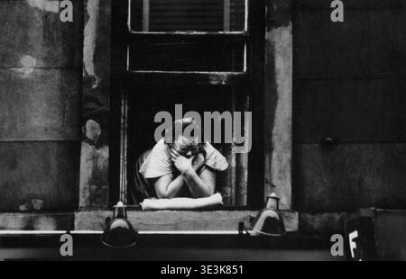 Dieses überzeugende Schwarz-weiß-Straßenfoto aus den 1950er Jahren, das wahrscheinlich von Angelo Rizzuto aufgenommen wurde, zeigt eine Frau, die sich mit überkreuzten Armen aus einem Fenster lehnt und auf der Fensterbank ruht. Ihre Brille und Haltung deuten auf einen Moment der Betrachtung oder Beobachtung des Alltags im alten New York hin. Stockfoto