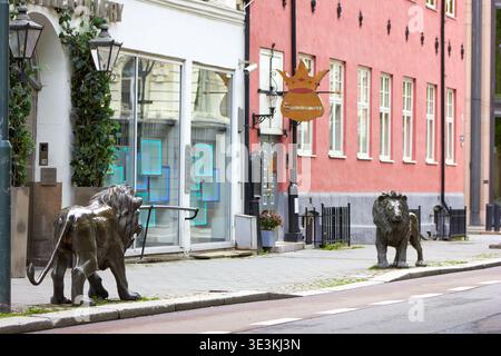 Zwei bronzene Löwenstatuen stehen auf einem Bürgersteig vor bunten Gebäuden. Auf der linken Seite zeigt ein weißes Gebäude mit großen Glasfenstern den Stockfoto