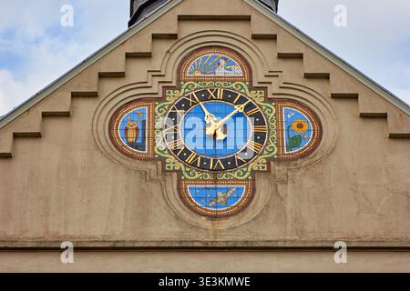 Aus nächster Nähe eine kunstvoll verzierte, farbenfrohe Uhr, die am Giebelende einer historischen Gebäudefassade montiert ist. Die zentrale Uhr ist mit hellblauen Fliesen versehen Stockfoto