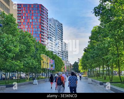 Ein von Bäumen gesäumter Fußgängerboulevard in einer modernen europäischen Stadt, in dem mehrere Personen vor der Kamera in Richtung eines modernen Hochhauses laufen Stockfoto