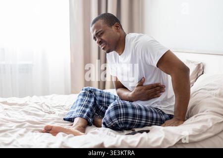 Junger schwarzer Mann, der auf dem Bett sitzt und den Magen mit Schmerzausdruck hält Stockfoto