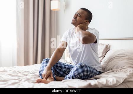 Schwarzer Mann, der auf dem Bett sitzt und den Hals vor Schmerzen hält, Muskelanstrengung hat Stockfoto