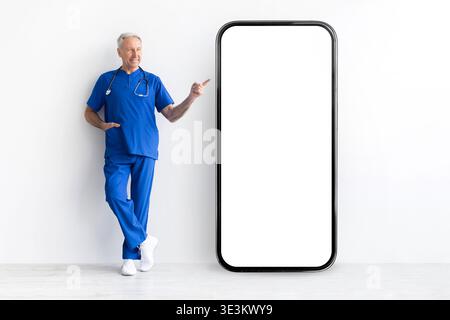 Leitender Arzt in blauen Schutten, der auf ein riesiges Smartphone mit leerem Bildschirm zeigt Stockfoto