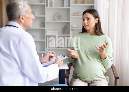 Schwangere Frau erklärt dem Arzt während der pränatalen Sprechstunde in der Klinik Symptome Stockfoto