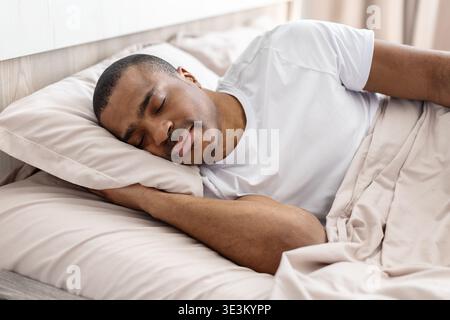 Hübscher junger schwarzer Mann, der friedlich auf dem Bett unter der Decke schläft Stockfoto