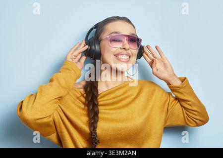Eine junge Frau trägt große Kopfhörer und lächelt, während sie Musik hört. Sie steht an einem sonnigen Tag vor einer hellblauen Wand und zeigt ihre Freude Stockfoto