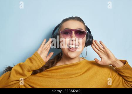 Eine junge Frau trägt Sonnenbrille und Kopfhörer, lächelt und genießt Musik. Sie hat einen fröhlichen Ausdruck und steht vor einer hellblauen Wand Stockfoto