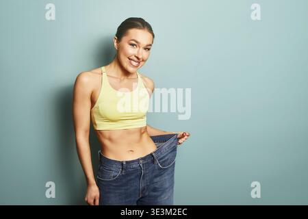 Eine Frau steht in einem Studio und hält ihre übergroße Jeans mit einem großen Lächeln hoch. Sie trägt ein gelbes Oberteil und sieht mit ihrer Leistung zufrieden aus. Die Ba Stockfoto