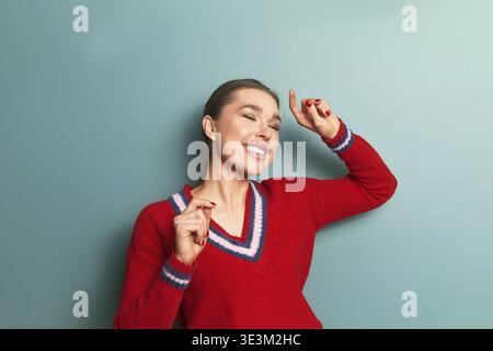 Eine Frau lächelt und tanzt in einem roten Pullover. Sie genießt Musik und drückt mit ihren Bewegungen Glück aus. Der Hintergrund ist ein durchgehend blauer Farbton Stockfoto