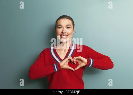 Eine Frau steht vor einer grünen Wand und macht mit ihren Händen eine Herzform. Sie lächelt weit und trägt einen roten Pullover mit Streifen. Die Einstellung ist Stockfoto