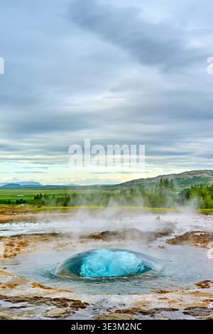 Strokkur-Geysir bricht aus und schickt Wasser hoch in die Luft. Die umliegende Landschaft zeigt Grün und ferne Berge. Stockfoto