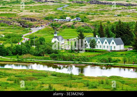 Touristen spazieren auf Wegen im Nationalpark Thingvellir. Grüne Felder und ein Fluss liegen in der Nähe einer kleinen Kirche und Häuser. Stockfoto