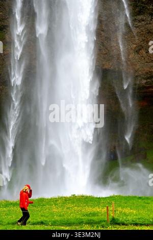 Eine Frau läuft in der Nähe des Seljalandsfoss Wasserfalls in Island. Das Wasser stürzt herunter, während sie die frische Luft und die Landschaft genießt. Stockfoto