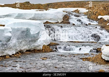 Wasser fließt über Felsen und Eis bei Djupvegur in Island. Schnee ist in der Nähe des Baches vorhanden. Die Szene zeigt die Natur in kaltem Klima. Stockfoto