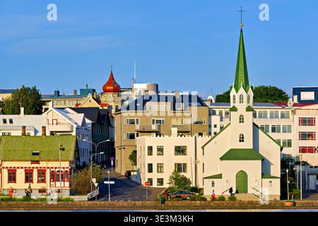 Die lutherische Freie Kirche steht am Tjornin See in Reykjavik. Menschen laufen in der Nähe, und Gebäude umgeben die Gegend unter klarem Himmel. Stockfoto