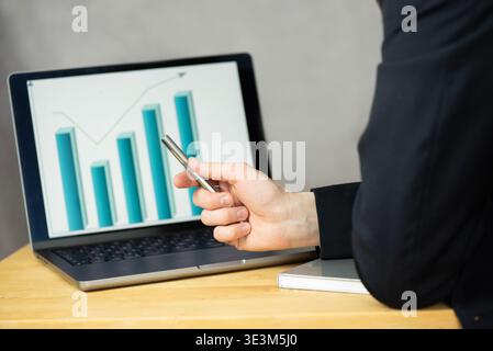 Ein Mann schaut auf einen Laptop, der ein Balkendiagramm anzeigt. Er hält einen Stift in seiner rechten Hand und zeigt auf das Diagramm. Der Laptop steht auf einem Tisch. Stockfoto