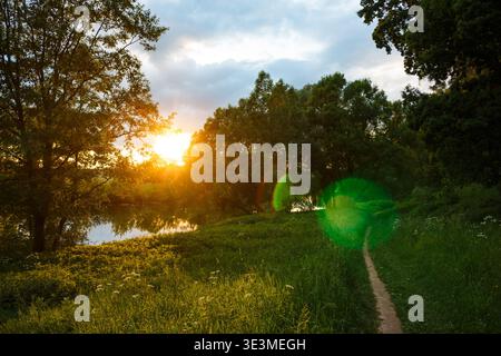 Herrliche Sonnenfackeln erleuchten einen gewundenen, unbefestigten Pfad durch lebhaftes Grün neben einem ruhigen reflektierenden Teich und schaffen einen magischen Abend-Vibe Stockfoto