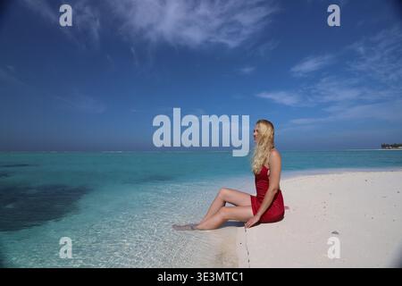 Junge blonde Frau in rotem Kleid sitzt auf weißem Sandbank-Rand mit Füßen in klarem türkisfarbenem Wasser, Malediven, Indischer Ozean Stockfoto